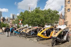 Mehrere Velotaxis mit Fahrer*innen warten in Frankfurt auf Fahrgäste, im Hintergrund die Skyline.