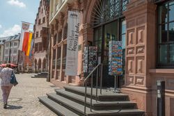 Eingang zur Tourist Information Römer. Ein großes Banner und zwei Postkartenständer stehen vor dem historischen Gebäude mit breiten Treppenstufen.