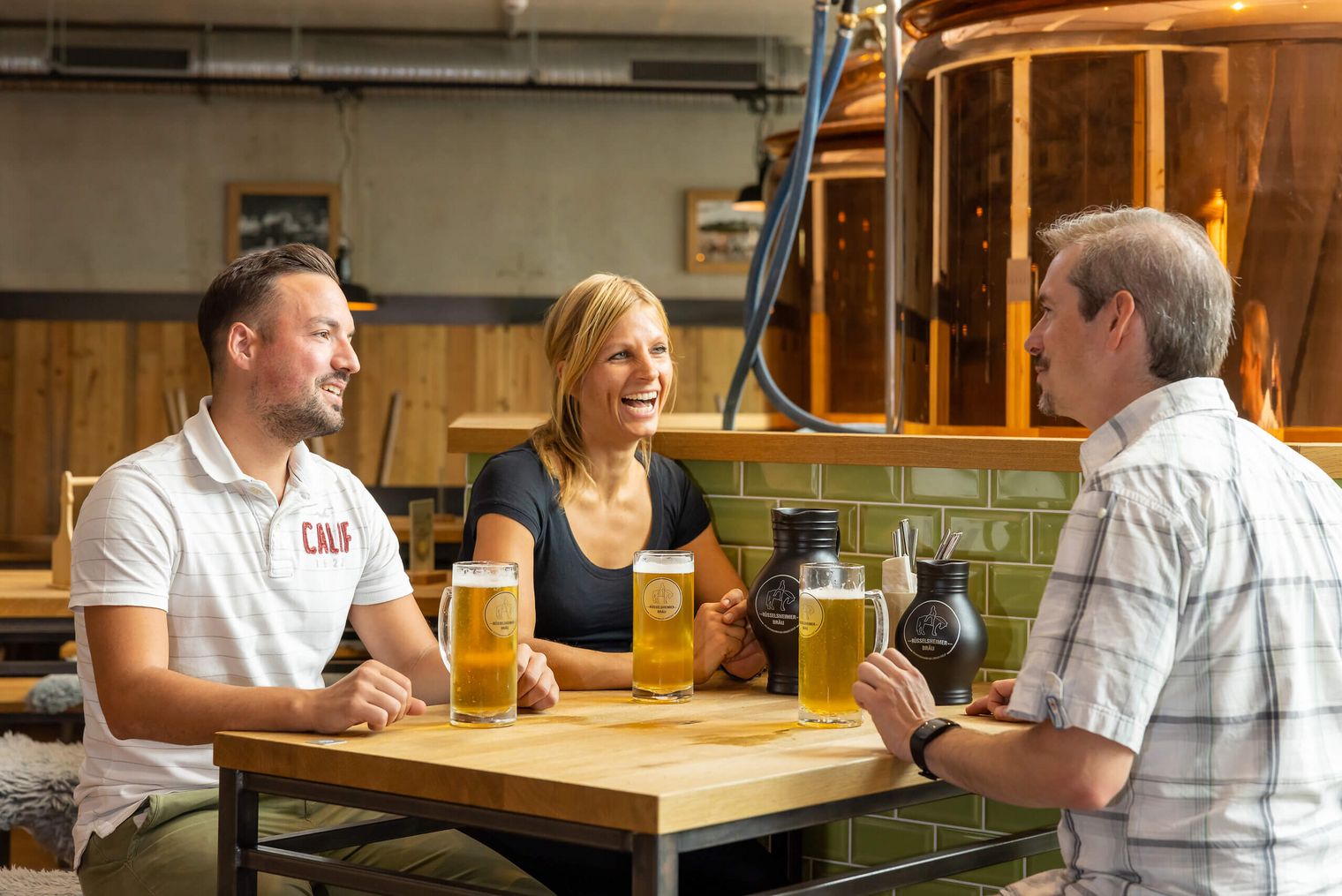 Drei Personen sitzen in der Hausbrauerei des MK Hotels in Rüsselsheim und genießen frisch gezapftes Bier.