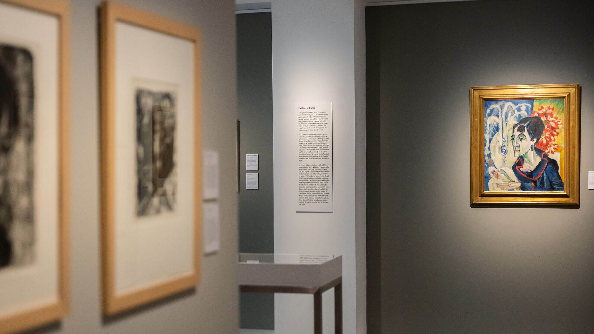 Blick in einen Ausstellungsraum im Kirchnerhaus Aschaffenburg mit gerahmten Kunstwerken an den Wänden und einem farbenfrohen Gemälde in einem goldenen Rahmen auf einer grauen Wand.