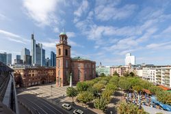 Außenansicht der Frankfurter Paulskirche aus rotem Sandstein, umgeben von Bäumen und einem großen Platz. Im Hintergrund ragen moderne Hochhäuser der Frankfurter Skyline in den Himmel, der von leichten Wolken bedeckt ist.