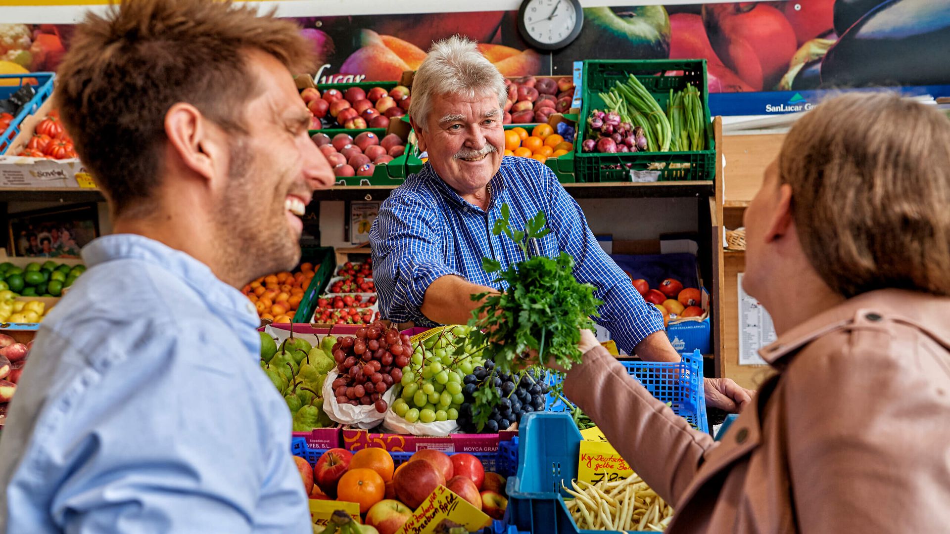 Ein Marktverkäufer in der Kleinmarkthalle Frankfurt reicht frische Kräuter, umgeben von buntem Obst und Gemüse sowie lachenden Kund*innen.