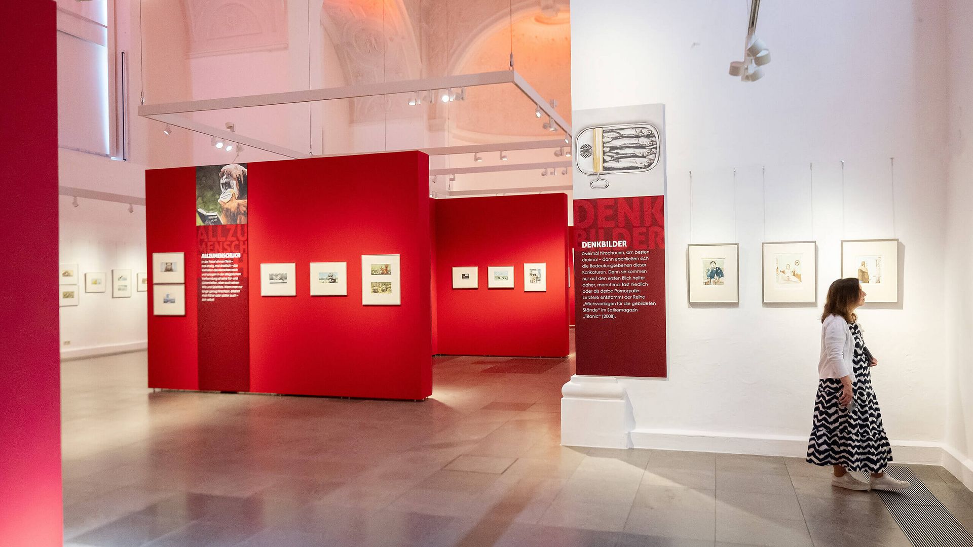Ausstellungsraum in der Kunsthalle Jesuitenkirche Aschaffenburg mit roten Wänden, gerahmten Zeichnungen und einer Besucherin, die die Kunstwerke betrachtet.