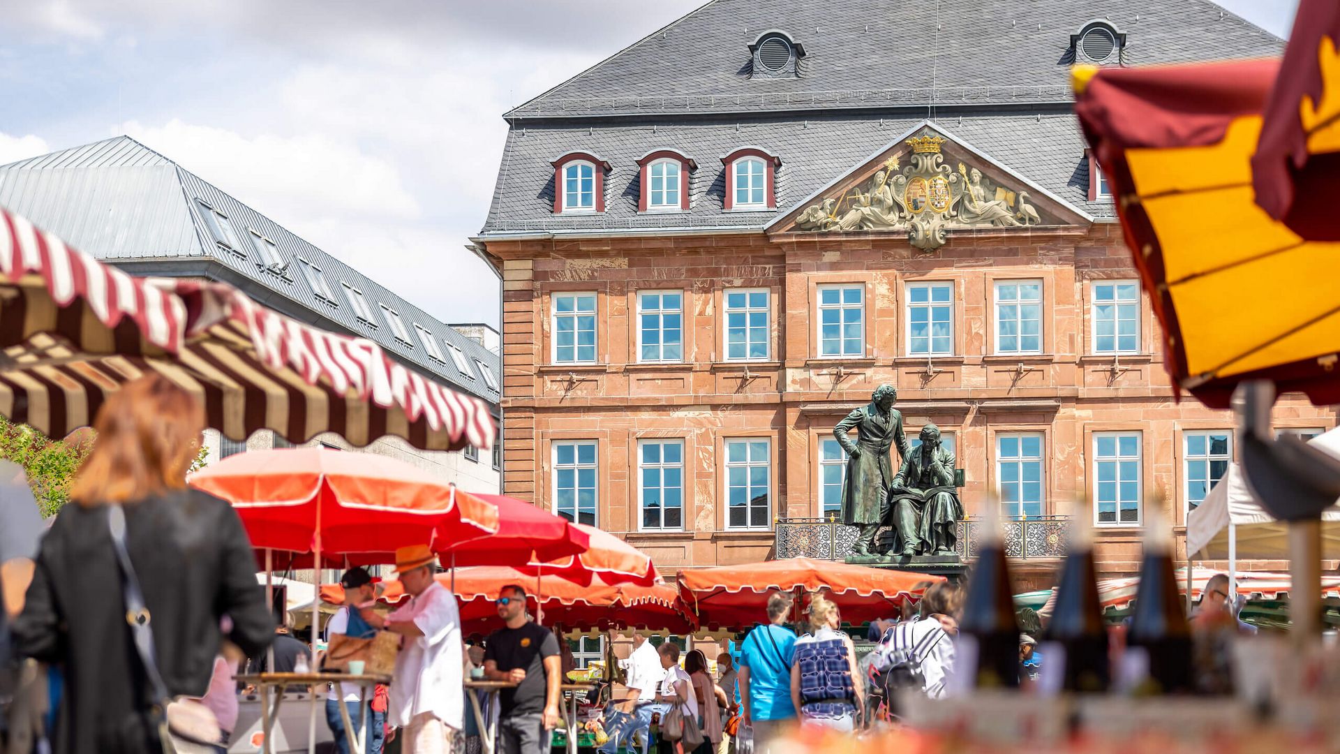 Marktszene mit roten Sonnenschirmen und Ständen. Im Hintergrund ist das Gebäude mit dem Brüder-Grimm-Denkmal zu sehen. Menschen flanieren zwischen den Ständen.