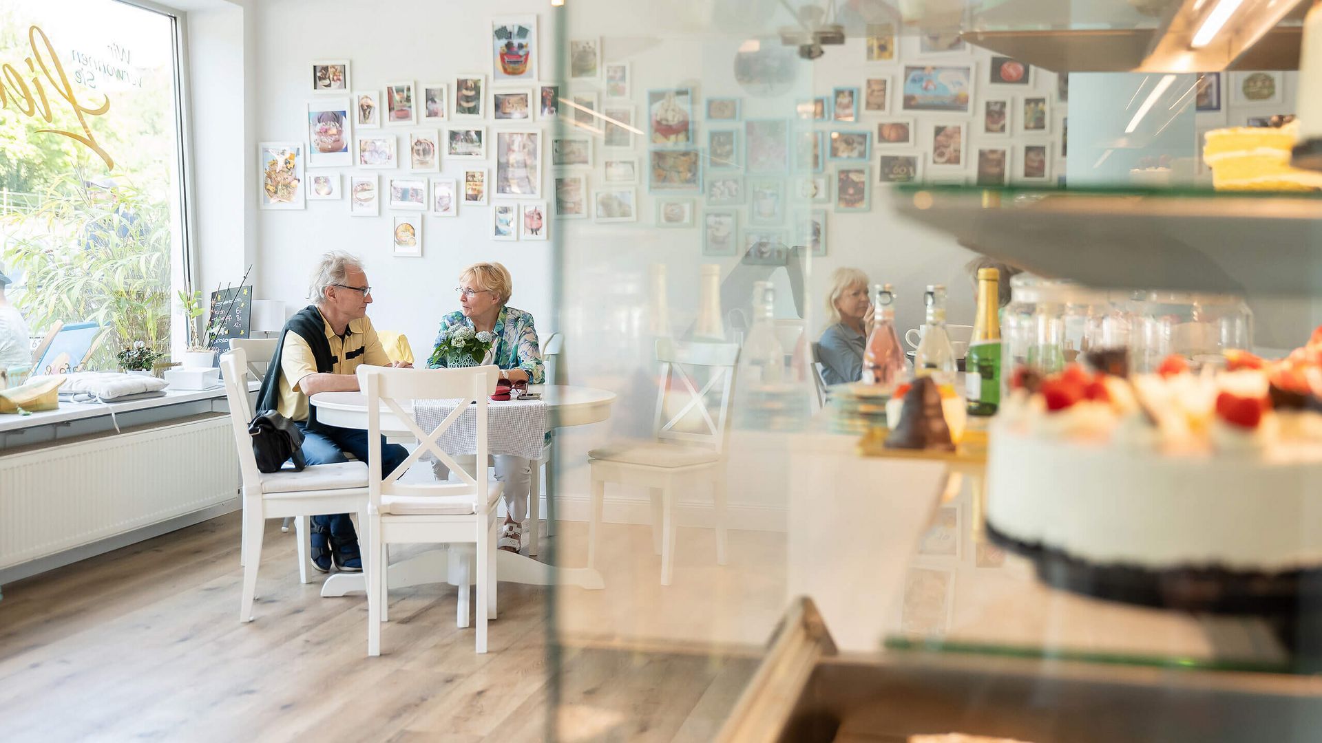 Helles und gemütliches Café mit Gästen an den Tischen und Kuchen in der Auslage im Vordergrund.