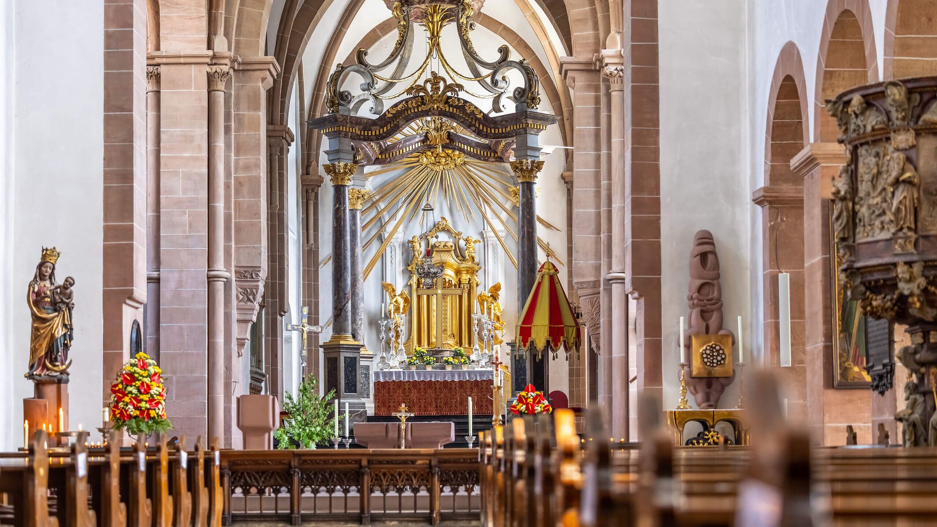Innenansicht der Stiftsbasilika Aschaffenburg mit prächtigem Altar, goldenen Verzierungen und Reihen aus Holzbankreihen.