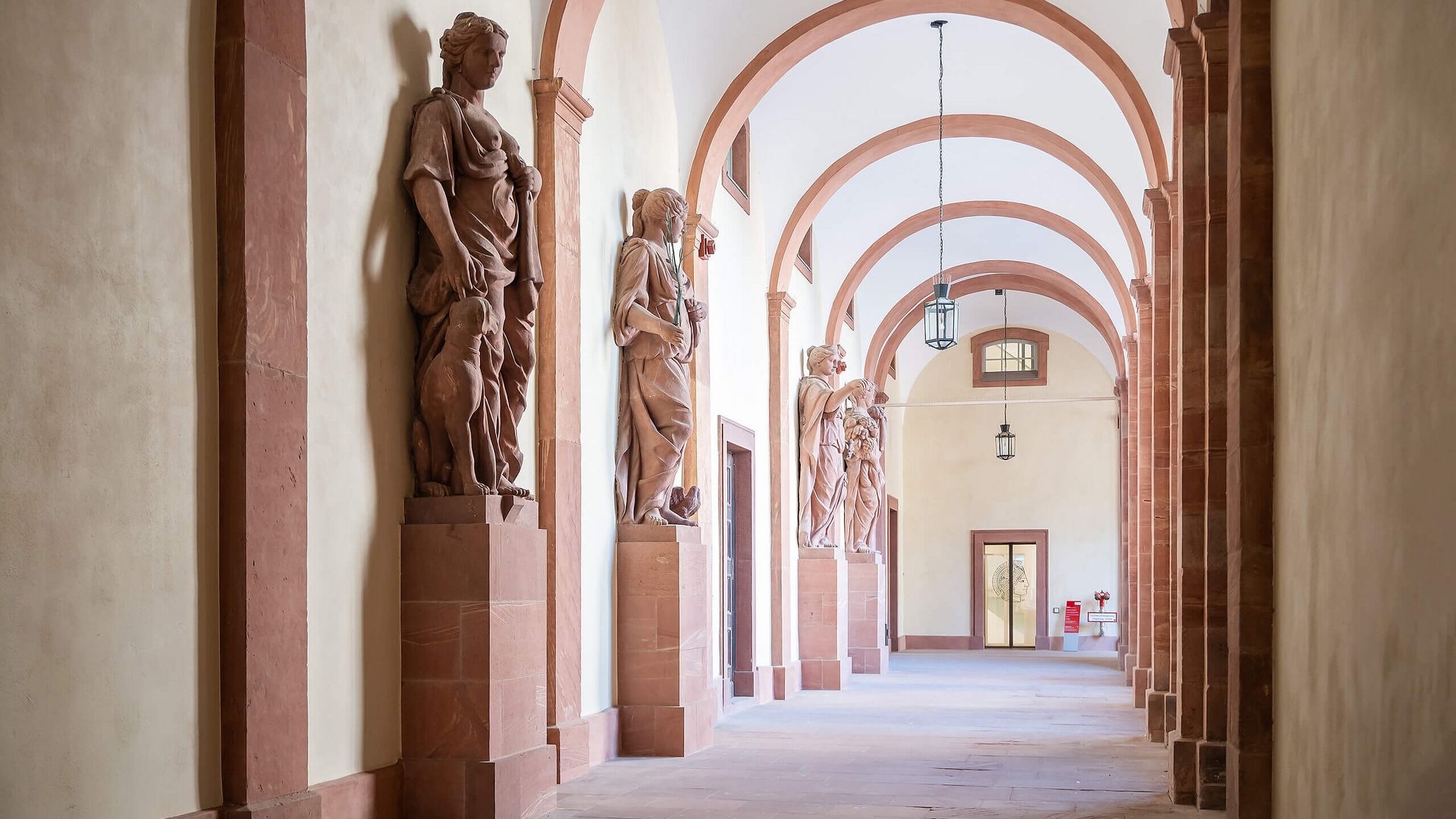 Säulengang im Residenzschloss Darmstadt mit Sandsteinfiguren entlang der Wand und Gewölbebögen.
