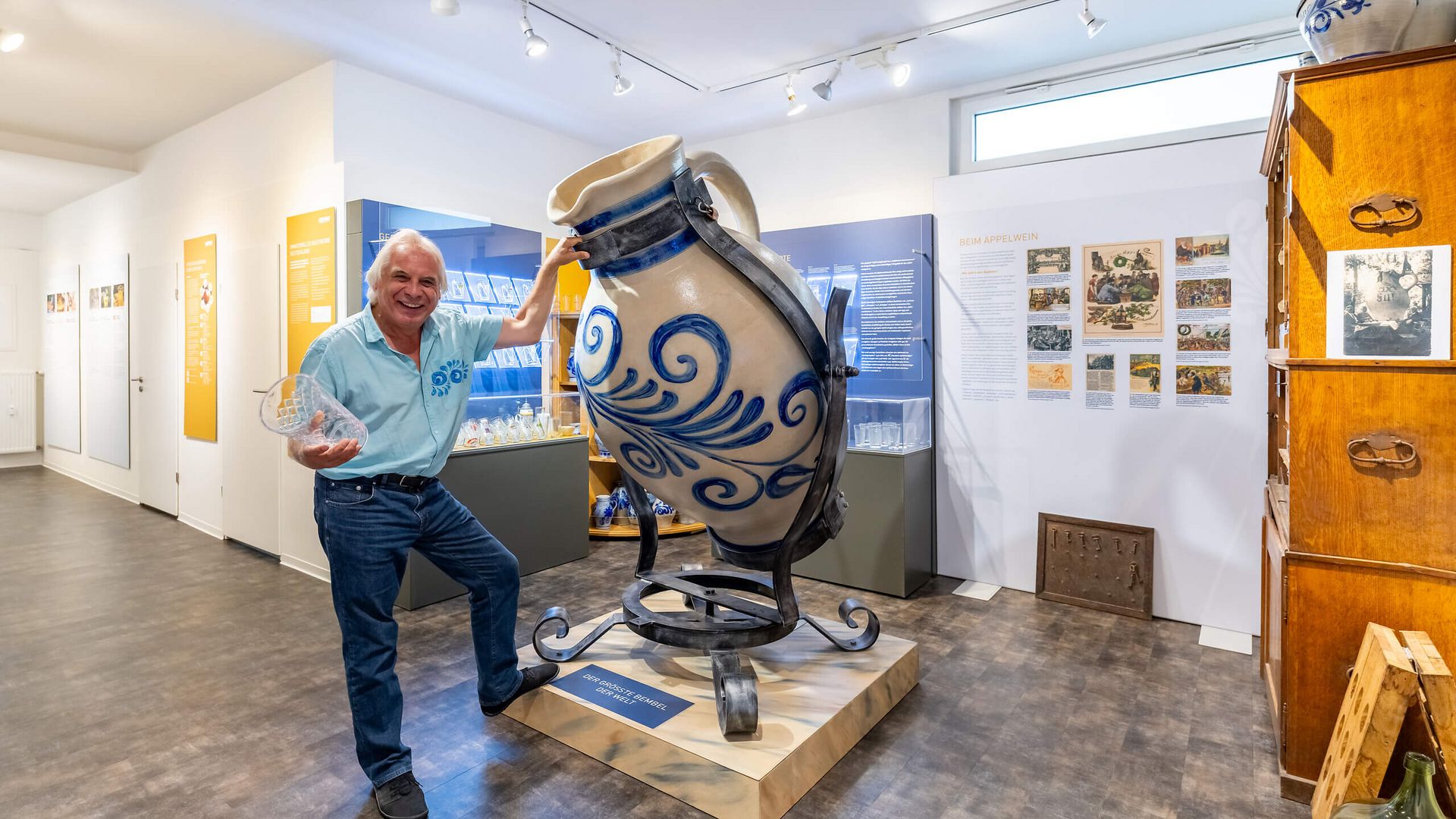 Ein Mann posiert lachend neben einem überdimensionalen Apfelwein-Krug im Geripptes Museum Frankfurt. Im Hintergrund sind Ausstellungswände und weitere Krüge zu sehen.