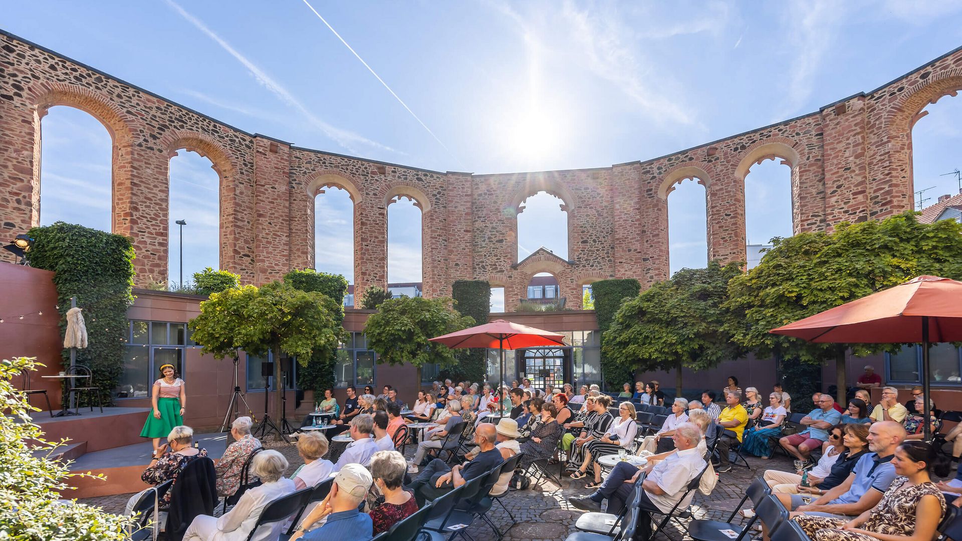 Publikum sitzt auf Stühlen in einem Innenhof mit den historischen Mauern der Wallonisch-Niederländischen Kirche. Eine Schauspielerin steht auf der Bühne, die Sonne scheint.