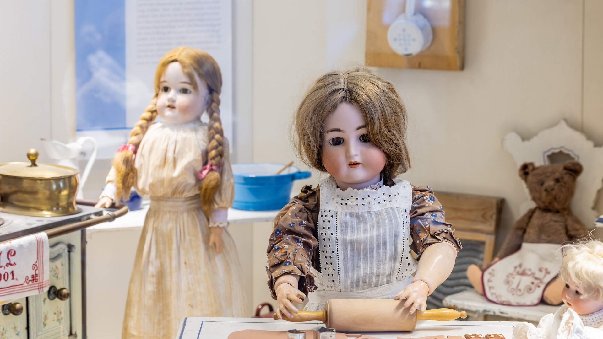 Zwei alte Porzellanpuppen in historischer Kleidung stehen in einer Puppenküche. Eine Puppe rollt Teig aus, während im Hintergrund ein Teddybär sitzt.